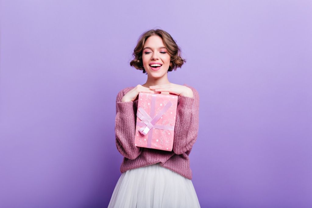 glamorous-girl-with-curly-short-hair-posing-with-pink-present-box-laughing-attractive-female-model-with-christmas-gift-isolated-purple-wall-smiling_197531-7950.jpg